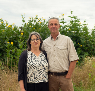 KI generiert: Das Bild zeigt ein Paar, das in einem Garten oder einer ländlichen Umgebung vor hohen, blühenden Pflanzen steht. Beide Personen lächeln leicht in die Kamera.