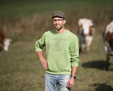 KI generiert: Ein Mann steht lächelnd auf einer Wiese, im Hintergrund befinden sich einige Kühe. Er trägt ein grünes Sweatshirt, eine Kappe und eine Brille.