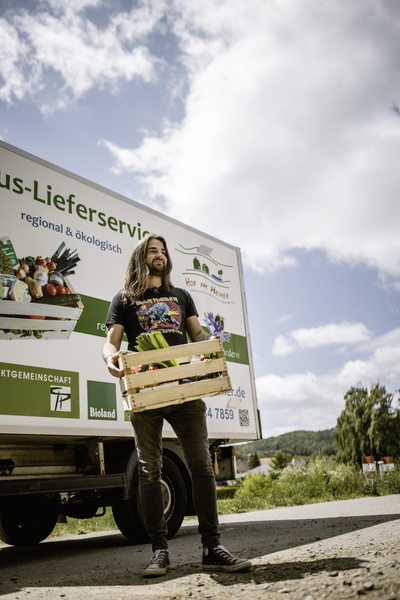 KI generiert: Ein Mann steht vor einem Lieferwagen und hält eine Gemüsekiste. Text: „regional & ökologisch“.